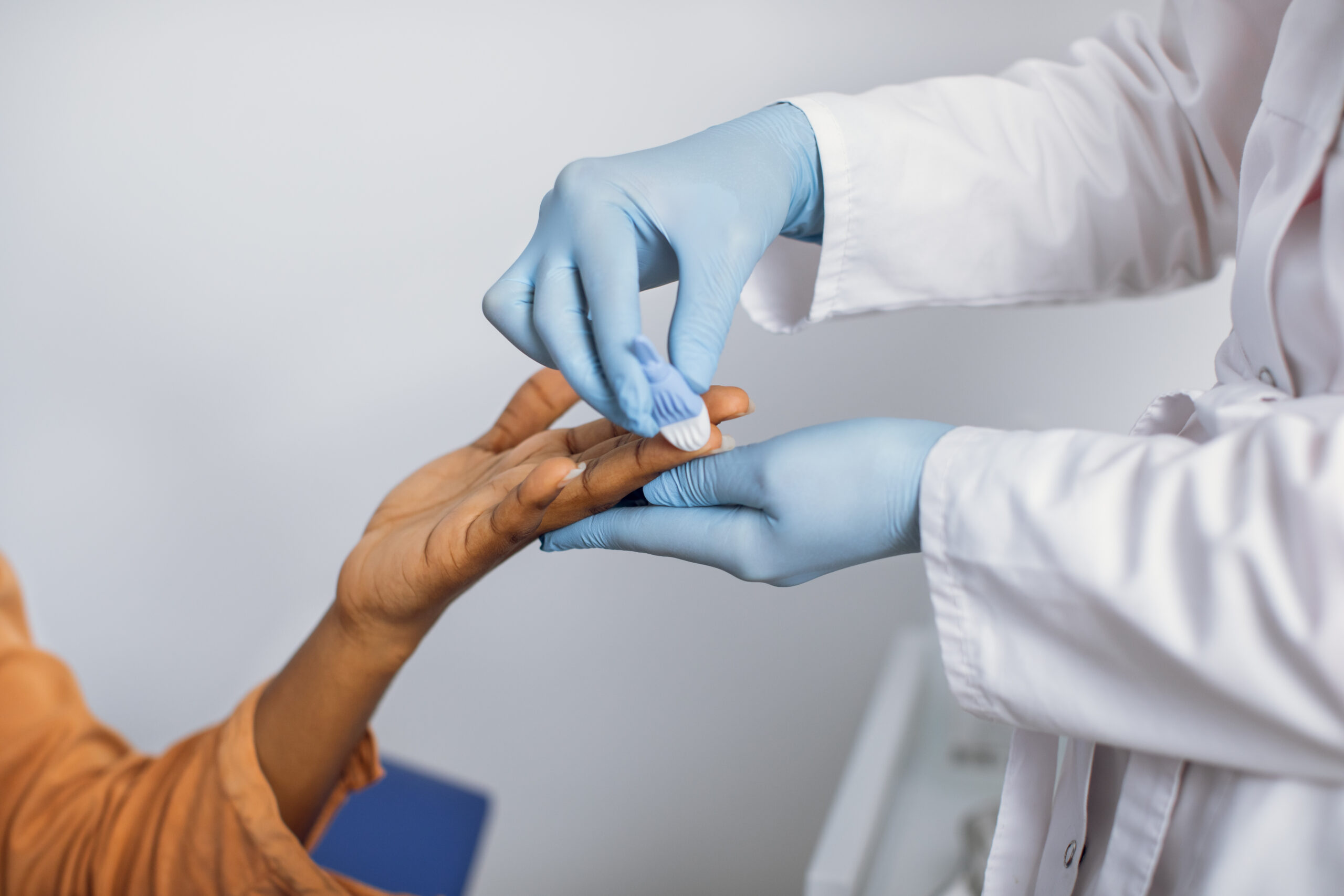 Close up photo of the hands of a female patient with an orange top and a worker in white coat and gloves, taking the patient's blood sample.